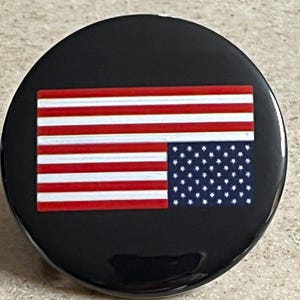 May include: A black button with a white circle in the center. The circle contains a red, white, and blue American flag with the stars and stripes reversed.