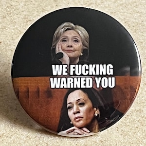 May include: A black and white button with a picture of Hillary Clinton and Kamala Harris. The text on the button reads "WE FUCKING WARNED YOU".
