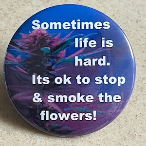May include: A round button with a blue and purple background featuring a cannabis bud graphic. White text reads: "Sometimes life is hard. Its ok to stop & smoke the flowers!"