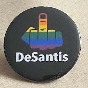 Könnte beinhalten: Ein schwarzer Knopf mit einem Regenbogen-Mittelfinger und dem Text "DeSantis" in Weiß.