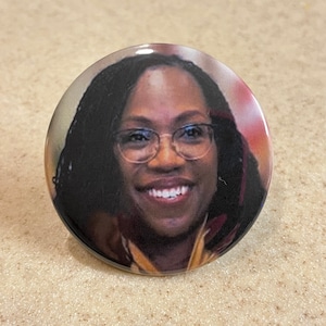 May include: A close-up of a button featuring a smiling woman with dark brown hair and glasses. She is wearing a red and gold shirt.