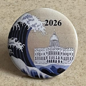 Puede incluir: Un botón con un gráfico de una gran ola rompiendo sobre el Capitolio de los Estados Unidos. La ola es azul y blanca, y el edificio es blanco. El año 2026 está impreso en negro en la parte superior del botón.