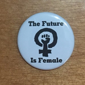 Puede incluir: Un botón blanco con texto negro y un símbolo feminista negro. El texto dice "The Future Is Female."