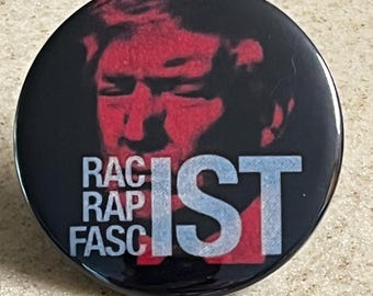 May include: A round, black button with a red image of a person's face and the words "RAC RAP FASC IST" in white and light blue. The button has a glossy finish.