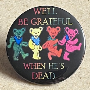 May include: A round, black button featuring four colorful dancing bear illustrations. The text "WE'LL BE GRATEFUL WHEN HE'S DEAD" is printed in rainbow colors around the bears. The bears are green, yellow, red, and pink.