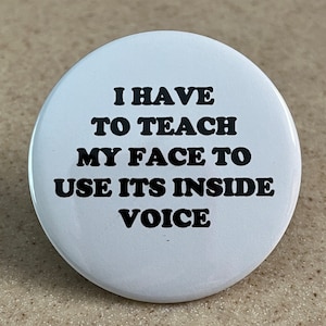 Puede incluir: Un botón redondo blanco con texto negro que dice "I HAVE TO TEACH MY FACE TO USE ITS INSIDE VOICE". El botón está sobre una superficie de color neutro.