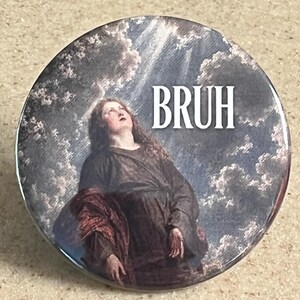 May include: A circular pin-back button featuring a classical painting of a person with long hair looking upward toward a cloudy sky. The word BRUH is printed in white, sans-serif capital letters in the upper right portion of the circular design.