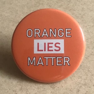 Puede incluir: Botón naranja con el texto "ORANGE LIES MATTER" en letras blancas. La palabra "LIES" está en un cuadro rojo.