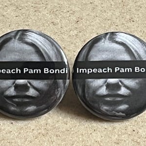 May include: Two round buttons with a black and white image of a person's face. A black horizontal bar across the face has the words "Impeach Pam Bondi" in white. The buttons are on a light brown surface.