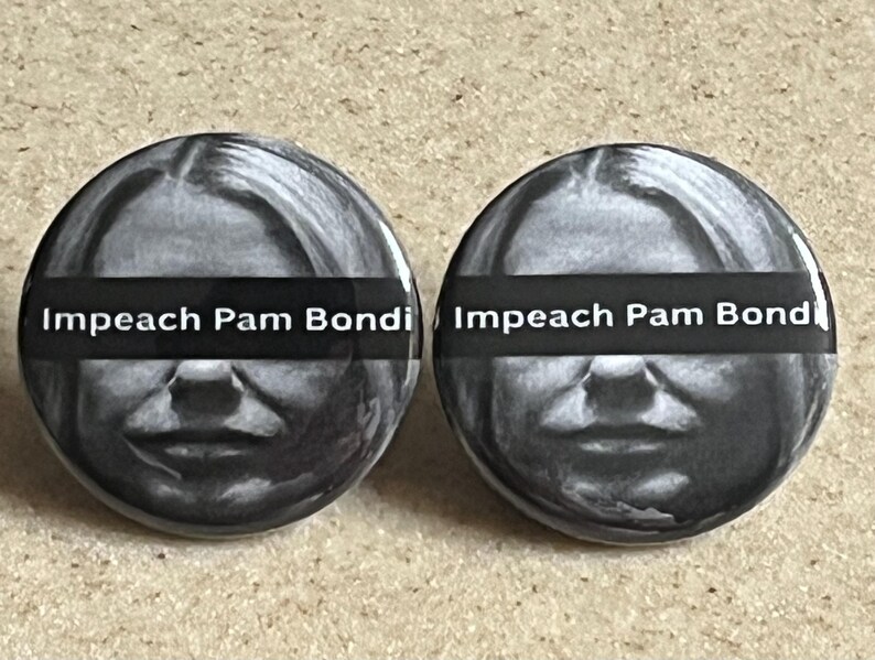May include: Two round buttons with a black and white image of a person's face. A black horizontal bar across the face has the words "Impeach Pam Bondi" in white. The buttons are on a light brown surface.