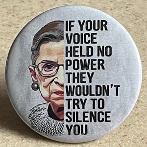 May include: A round button with a portrait of Ruth Bader Ginsburg on the left and the quote "IF YOUR VOICE HELD NO POWER THEY WOULDN'T TRY TO SILENCE YOU" in black text on the right. The button has a light gray background.