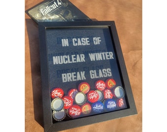 May include: A black framed picture with the text "IN CASE OF NUCLEAR WINTER BREAK GLASS" and a collection of red, blue, and white bottle caps with the text "Nuka Cola" inside the frame.