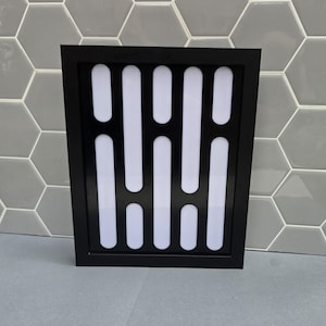 May include: Black framed wall art with a white and black geometric pattern. The artwork displays vertical white rectangles on a black backdrop. The frame has a simple, modern design.