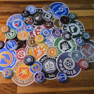 May include: A selection of colourful round stickers and bottle caps featuring diverse designs and text, such as "Electric", "Double Tap", and "Real Beer". The stickers and caps are displayed on a wooden surface.