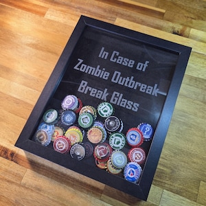 Puede incluir: Un marco con un marco negro, lleno de coloridas tapas de botellas. El texto "In Case of Zombie Outbreak - Break Glass" está impreso en el fondo oscuro. Las tapas de botellas tienen varios diseños y colores.