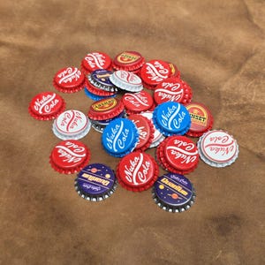 May include: A collection of metal bottle caps featuring various designs, including red caps with white Nuka Cola text, blue caps with white Nuka Cola text, and dark purple caps with yellow Quantum text and planet graphics, alongside yellow Sunset caps.