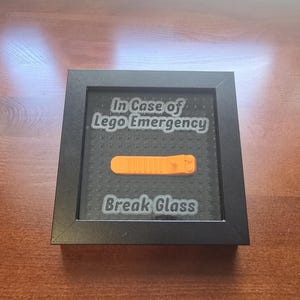 May include: A framed piece with the text "In Case of Lego Emergency" and "Break Glass". Inside the frame is an orange Lego brick separator. The frame is black and sits on a wooden surface.