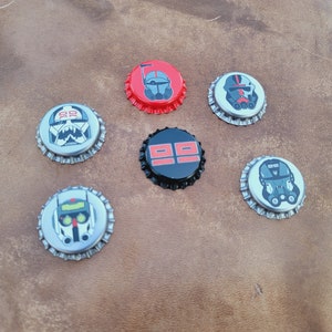 Pode incluir: Seis tampas de garrafa com diferentes desenhos. As tampas são prateadas com desenhos pretos e vermelhos. Uma tampa tem um fundo preto com números vermelhos 99. Outra tampa tem um fundo vermelho com um desenho preto e branco de um capacete.