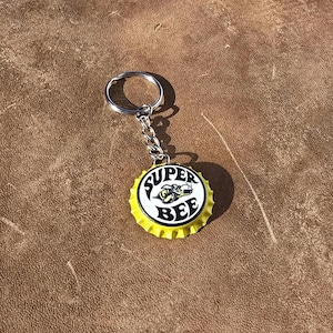 May include: A keychain featuring a yellow bottle cap with the words "SUPER BEE" in black and white, and a graphic of a bee. The keychain has a silver chain and ring.