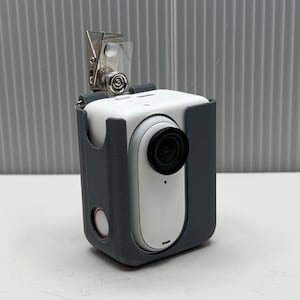 May include: A white and gray body camera with a black lens and a red button. The camera is attached to a gray case with a clip.