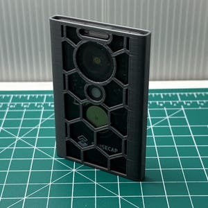 Peut inclure: Un boîtier de protection rectangulaire gris foncé avec un motif de grille hexagonale en relief sur le devant. Le boîtier présente des découpes pour un objectif d'appareil photo et un capteur, avec le texte SENSECAP imprimé en blanc en bas.
