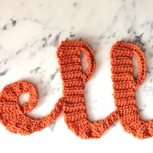 May include: A crocheted "hello" sign in a warm orange hue. The word is crafted from textured yarn, with each letter carefully formed. The sign is designed to be a decorative accent.