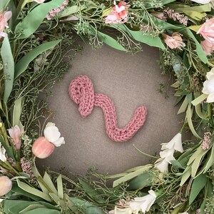 May include: A pink crocheted letter S surrounded by a wreath of green leaves and pink flowers.