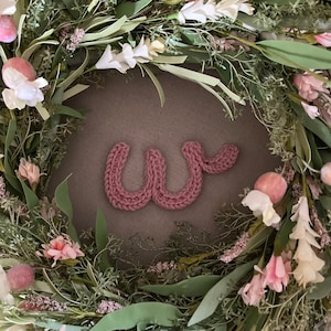 Peut inclure: Une lettre W en tricot rose est affichée sur un fond brun entouré d'une couronne florale verte avec des fleurs roses et blanches.