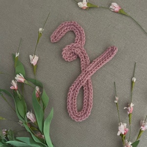 May include: A pink knitted letter 'Y' on a gray background. The letter is made of yarn and is a decorative item.