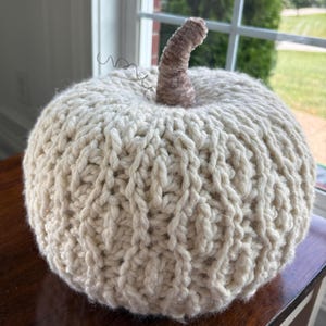 May include: A round, cream-colored knitted pumpkin decoration featuring a thick, textured cable knit pattern. The pumpkin is topped with a short, curved, light brown stem made of wrapped yarn and a thin, dark, curled wire tendril.