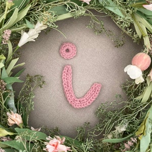 May include: A pink knitted letter "i" with a pink knitted dot on top, surrounded by a green floral wreath.