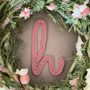 May include: A pink knitted letter "h" surrounded by a green floral wreath with pink and white flowers.