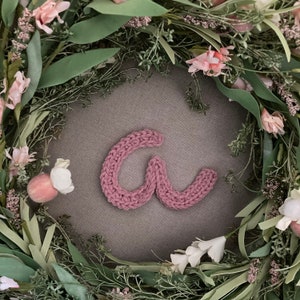 May include: A knitted, mauve-colored letter "a" rests on a gray surface, surrounded by a wreath of greenery, white and pink flowers. The wreath is composed of various leaves and blossoms, creating a natural, decorative frame.