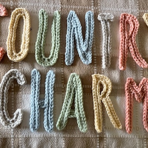 May include: Crocheted letters spelling "COUNTRY CHARM" in various pastel colors, including pink, yellow, green, and blue. The letters are arranged on a textured, neutral-toned fabric, creating a rustic, handmade aesthetic.