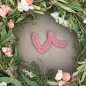 Puede incluir: Una letra "V" de punto de color rosa rodeada de una corona de hojas verdes y flores blancas y rosas.