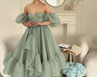 May include: A sage green off-the-shoulder gown featuring a structured sweetheart corset bodice, voluminous sheer puff sleeves, and a floor-length skirt with multiple layers of ruffled tulle fabric that create a full, textured silhouette.