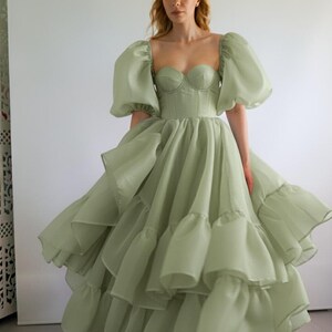 May include: A sage green midi dress featuring a structured corset-style bodice with a sweetheart neckline and voluminous puff sleeves. The skirt is composed of multiple layers of sheer, ruffled fabric that create a full, flared silhouette.