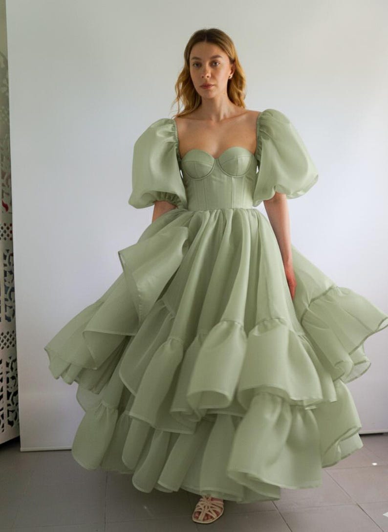 May include: A sage green midi dress featuring a structured corset-style bodice with a sweetheart neckline and voluminous puff sleeves. The skirt is composed of multiple layers of sheer, ruffled fabric that create a full, flared silhouette.