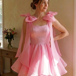 May include: A light pink mini dress featuring a square neckline, a fitted bodice, and a flared skirt with multiple layers. The dress includes two large, sheer organza bows on each shoulder with long, flowing ribbons extending down the front.