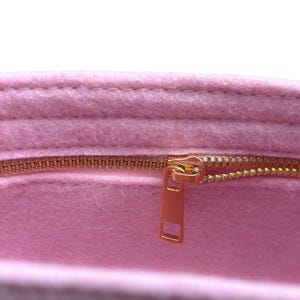Op de afbeelding: Close-up van een roze vilten organizer met een goudkleurige rits. De rits is gedeeltelijk open en onthult de binnenkant van de organizer. Het vilt heeft een zacht, getextureerd uiterlijk.