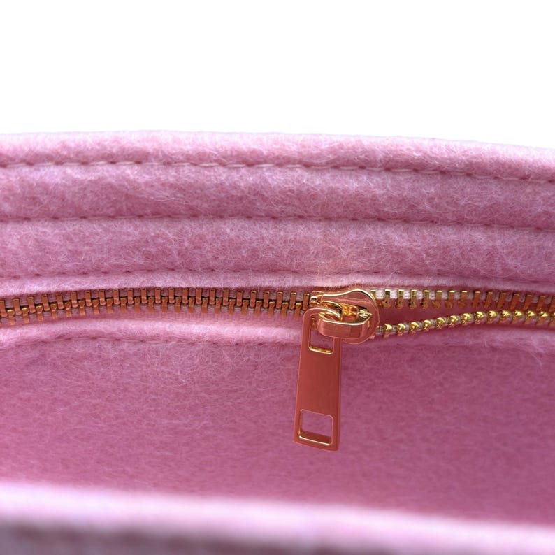 Op de afbeelding: Close-up van een roze vilten organizer met een goudkleurige rits. De rits is gedeeltelijk open en onthult de binnenkant van de organizer. Het vilt heeft een zacht, getextureerd uiterlijk.