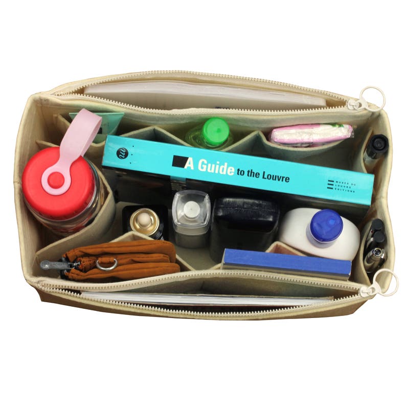 Puede incluir: Bolso organizador beige con cremallera y m&uacute;ltiples compartimentos, lleno de diversos art&iacute;culos, incluyendo un libro titulado "A Guide to the Louvre", una botella de agua, una peque&ntilde;a botella de loci&oacute;n, un cuaderno y bol&iacute;grafos.