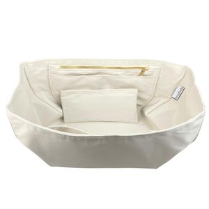 Könnte beinhalten: Cremefarbener Innentaschen-Organizer mit mehreren Taschen und einem Reißverschlussfach. Der Organizer besteht aus einem glatten, hellen Stoff und enthält einen kleinen, rechteckigen Beutel. Ein goldfarbener Reißverschluss ist sichtbar.