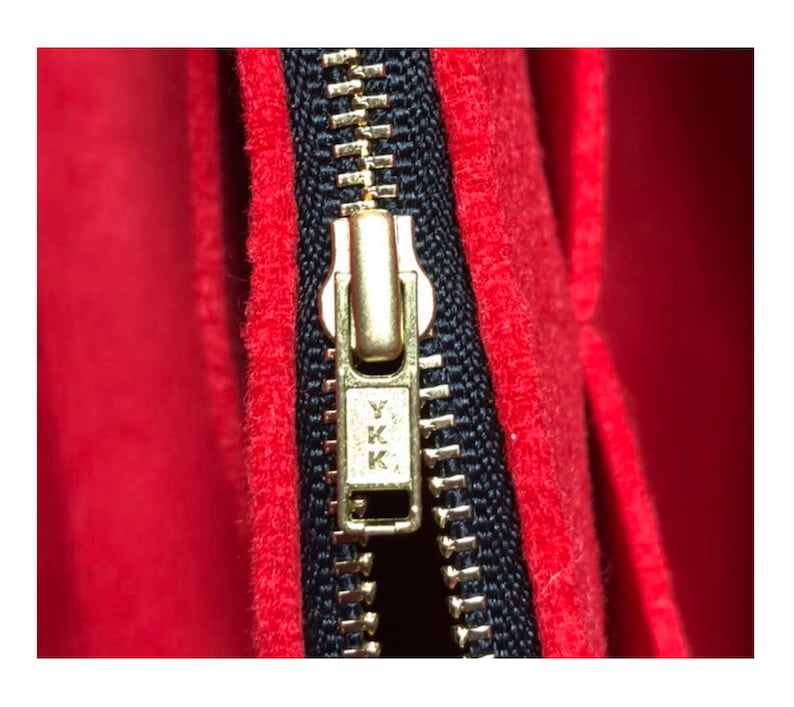 Pode incluir: Close-up de um z&iacute;per dourado com dentes pretos e um fundo de tecido vermelho. O puxador do z&iacute;per tem o texto "YKK" gravado.