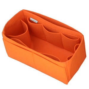 May include: A bright orange felt handbag organizer insert with a rectangular shape. The interior features multiple open compartments and dividers for storage. A small white fabric tag with black text is sewn onto the inner side wall.