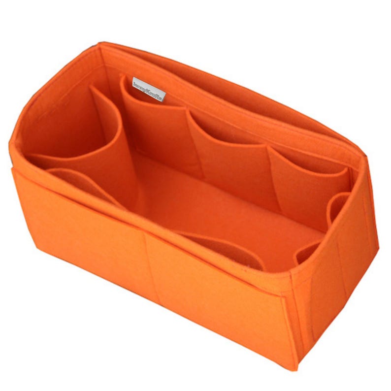 May include: A bright orange felt handbag organizer insert with a rectangular shape. The interior features multiple open compartments and dividers for storage. A small white fabric tag with black text is sewn onto the inner side wall.