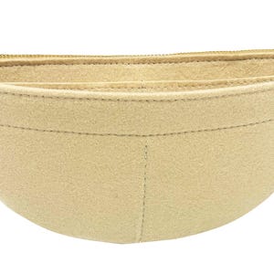 May include: A beige felt handbag organizer with a curved shape and a gold zipper closure. The organizer has multiple compartments and a small tag that reads "jennyKrafts".