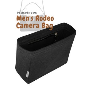 Peut inclure: Organisateur de sac photo en feutre noir avec une fermeture éclair sur le dessus. Conçu pour le sac photo Men's Rodeo. Le texte "DESIGNED FOR Men's Rodeo Camera Bag" est imprimé en haut.