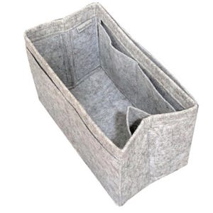 May include: A light gray felt bag organizer with a rectangular shape and structured walls. The interior features multiple compartments and dividers for storage. A small white fabric tag with the text "JennyKraft" is sewn onto the inner rim.