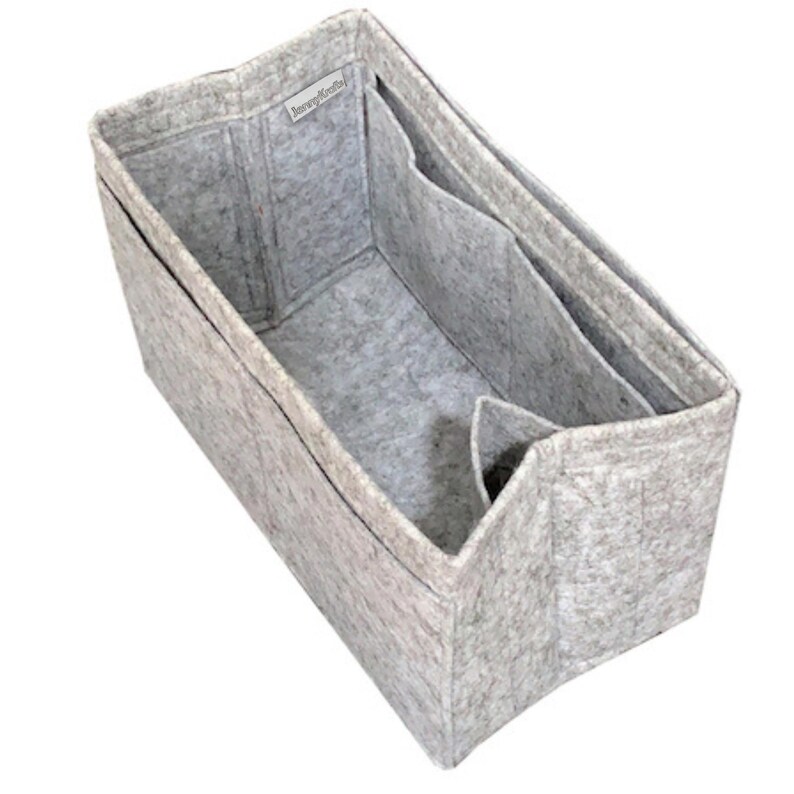 May include: A light gray felt bag organizer with a rectangular shape and structured walls. The interior features multiple compartments and dividers for storage. A small white fabric tag with the text "JennyKraft" is sewn onto the inner rim.